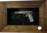 Engraved silver and black pistol displayed horizontally inside a dark wood frame with a black background and a circular white knob at the bottom right corner.