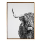 Black and white close-up of a Highland cow facing forward, showing one prominent curved horn and shaggy fur partially covering its eyes, framed in light wood.