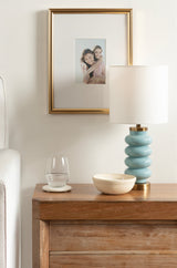 Curved light blue ceramic table lamp with brass accents and white shade on wooden bedside table, accompanied by a glass of water and small wooden bowl, under a gold-framed photo.