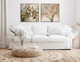 Minimalist living room with a white three-seat sofa beneath two framed nature paintings of olives and a tree, accompanied by a round woven coffee table and natural decor.