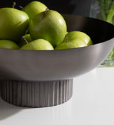Dark metal bowl with ribbed cylindrical base holding several green apples on a white surface, with a glass vase partially visible in the background.