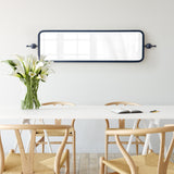 Rectangular Kasota mirror with thin black metal frame mounted horizontally above a white table, with wooden chairs and a clear vase of white calla lilies.