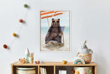 Framed canvas art of a bear wearing orange sunglasses sitting on a striped beach chair under an orange and white umbrella on a sandy beach.