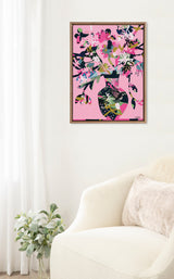 Abstract floral artwork with pink background featuring colorful overlapping shapes in dark green, blue, white, and pink, framed and hanging on a white wall above a cream chair.