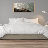 Framed canvas of a lighthouse on rocky coast overlooking sparkling ocean, hung above bed with white bedding in a minimalist gray bedroom.