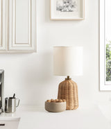 Table lamp with a ribbed, honey-colored rattan base and a white cylindrical linen shade on a clean white surface with a beige bowl of nuts beside it.