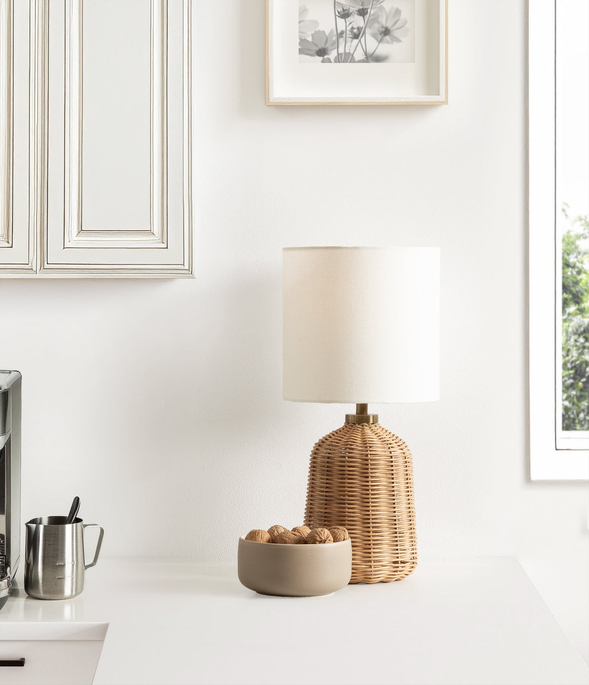 Table lamp with a ribbed, honey-colored rattan base and a white cylindrical linen shade on a clean white surface with a beige bowl of nuts beside it.