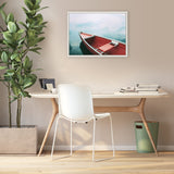 Framed canvas on beige wall depicting a red and white canoe floating on calm blue water, above a minimalist wooden desk with white chair and green plant.