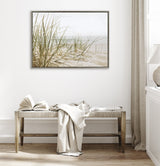 Framed canvas showing beach grass on sandy dunes with soft neutral tones, hanging above a wooden bench with a draped light fabric in a bright, minimalist room.