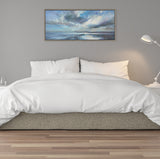 Horizontal framed canvas above bed featuring a seascape with blue-gray clouds and reflective water under a dynamic sky, set in a minimalist bedroom with white bedding.
