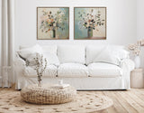 Minimalist living room with white slipcovered sofa, round woven ottoman, beige textured rug, and two framed botanical vase paintings in pastel tones on white wall.