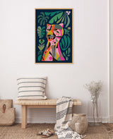 Framed Sylvie Leopard canvas with colorful abstract leopard and green foliage on dark blue background, hanging above a wooden bench with striped cushion and throw in a minimalist room.