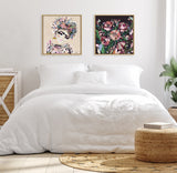 Minimalist bedroom with white bedding, framed colorful floral and abstract portraits above the bed, natural-toned woven ottoman and rug, and a side table with a potted plant.
