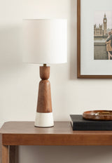 Table lamp with tapered wood and white ceramic base, topped with a simple white cylindrical lampshade, placed on a wooden console table next to a book and ashtray.