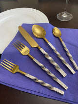 Set of gold flatware with textured silver twisted handles arranged on a purple cloth, beside a white plate and a wine glass on a dark wooden surface.