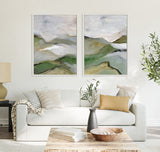 Two framed abstract landscape paintings with soft gray, green, and beige tones hung above a white sofa with neutral and yellow pillows in a modern living room.