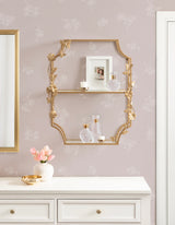 Gold metal tiered shelf with ornate floral accents holding decorative bottles and a framed photo against pale pink floral wallpaper above a white dresser with gold-handled drawers.