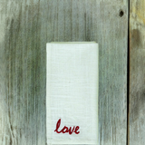 Folded white cloth napkin with textured fabric and red embroidered word 'love' in cursive, placed on weathered gray wooden surface with visible grain and knot.
