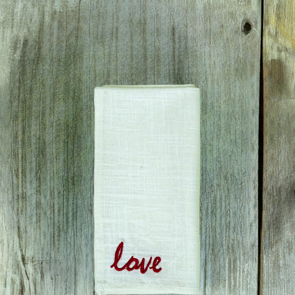 Folded white cloth napkin with textured fabric and red embroidered word 'love' in cursive, placed on weathered gray wooden surface with visible grain and knot.