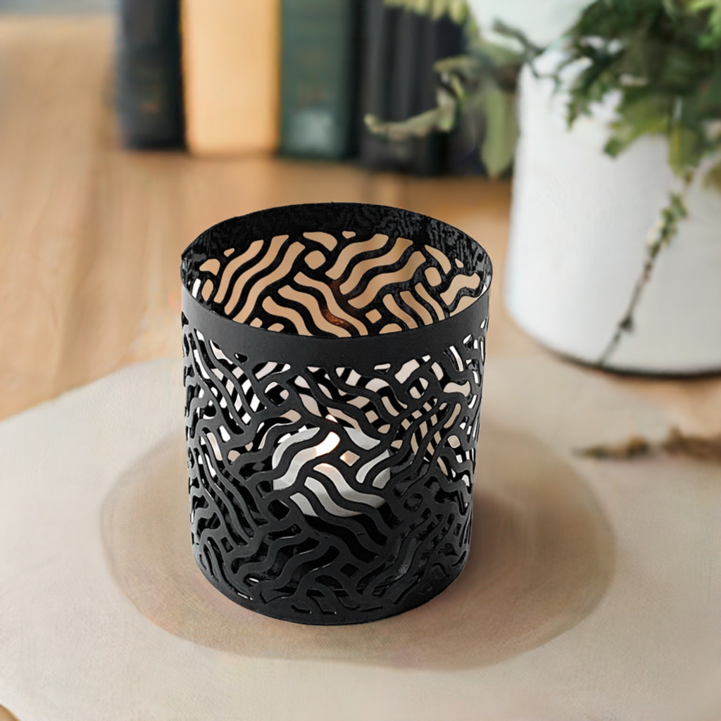 Black metal votive candle holder with an abstract wavy cutout pattern, glowing softly from a lit candle inside, placed on a light wood surface.