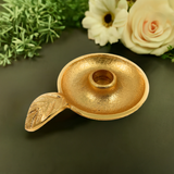 Round gold-toned metal candlestick holder dish with textured surface and leaf-detail handle, set against a black background with white and peach flowers and greenery
