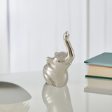 Small silver-finished elephant figurine with raised trunk on a reflective white surface next to a stack of two books with green and beige covers.