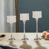 Set of three silver metal candle holders with square tops encrusted with clear crystal beads, varying in height, arranged on a light wooden surface with flowers nearby.