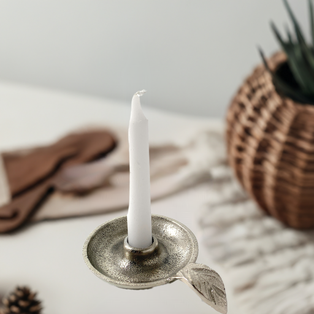 Silver-colored metal taper candlestick holder featuring a textured dish and a small leaf-shaped handle, holding a white unlit candle, on a neutral surface with soft-focus background items.