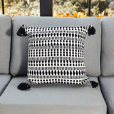 Square black and white patterned throw pillow with geometric shapes and black tassels on each corner, placed on a light gray couch seat with back cushions.