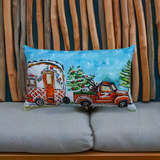 Rectangular pillow with hand-illustrated vintage truck towing a decorated caravan and Christmas tree, set against a blue sky, resting on a gray cushion with wooden sticks background.