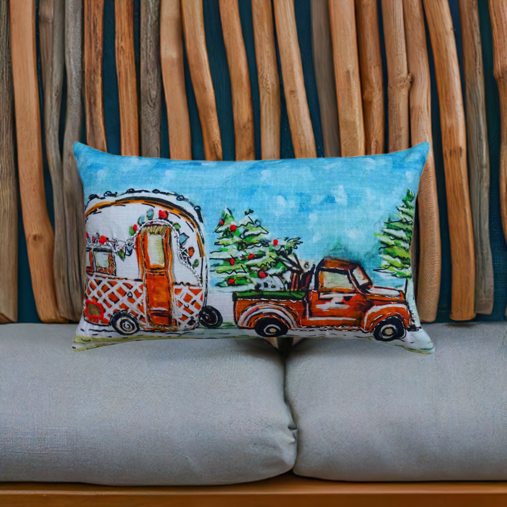Rectangular pillow with hand-illustrated vintage truck towing a decorated caravan and Christmas tree, set against a blue sky, resting on a gray cushion with wooden sticks background.