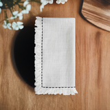 White linen napkin with black embroidered border and fringed edges, neatly folded on a warm wood surface next to a hexagonal wooden tray and blurred white flowers.