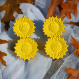Set of four bright yellow sunflower-shaped napkin rings with textured centers, arranged on a white fabric background with scattered brown autumn leaves.