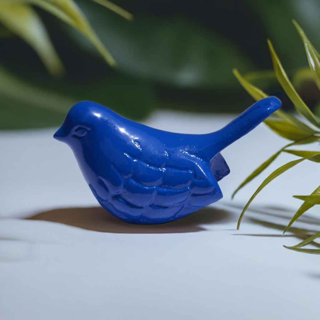 Small blue bird figurine with carved feather patterns and a smooth, rounded tail, set against a soft background with green plant leaves in the foreground.