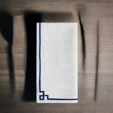 White linen napkin with a simple dark blue embroidered border featuring a corner square detail, shown folded on a textured wooden surface with shadows.