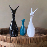 Three matte-finish ceramic deer figures in black, teal, and white colors standing on a round woven basket with a natural beige background.