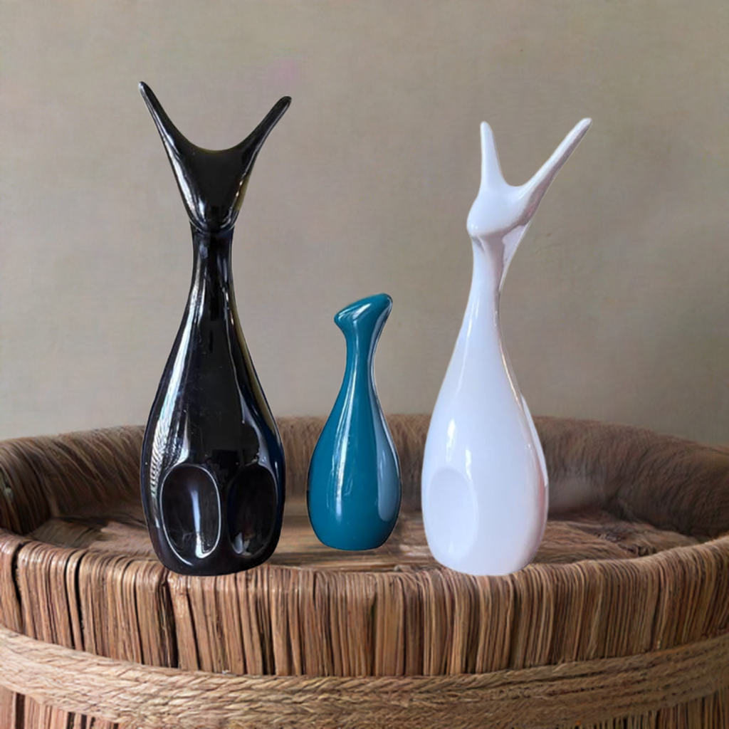 Three matte-finish ceramic deer figures in black, teal, and white colors standing on a round woven basket with a natural beige background.