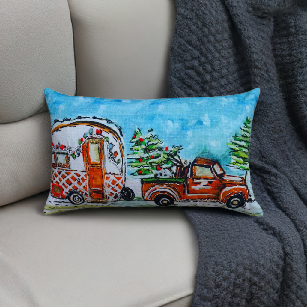 Rectangular pillow with hand-illustrated design of a vintage red truck carrying a Christmas tree and towing a decorated camper against a bright blue sky background.