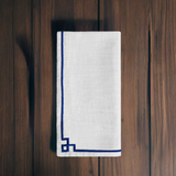 White cloth napkin with a simple blue embroidered border featuring a geometric corner knot, displayed folded on a dark wooden surface.