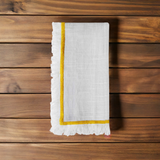 White linen napkin with a yellow embroidered border and fringed edges folded on a wooden surface with natural grain patterns.
