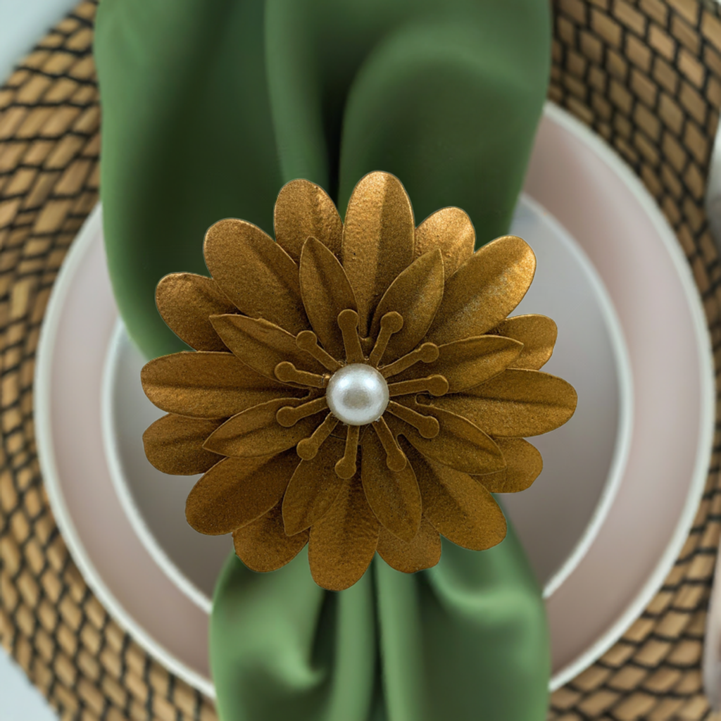 Golden floral napkin ring with layered petals and a central pearl, holding a neatly folded green napkin over a white plate on a woven placemat.