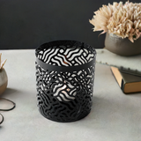 Black cylindrical votive candle holder with a wavy, abstract cutout pattern revealing a lit candle inside, placed on a light surface.