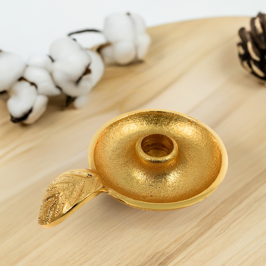 Round golden taper candlestick holder dish with a textured surface and a small attached leaf detail, placed on a light wood surface with cotton and pinecone decor.