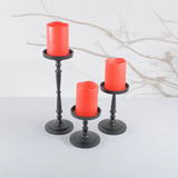 Set of three black metal pillar candle holders in varying heights holding red pillar candles, against a white background with faint brown twig shadows.