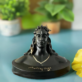 Black bust statue of Adiyogi with detailed facial features, hair styled with a crown, and 'Adiyogi' inscribed at the base, set against a blurred indoor background with plants.