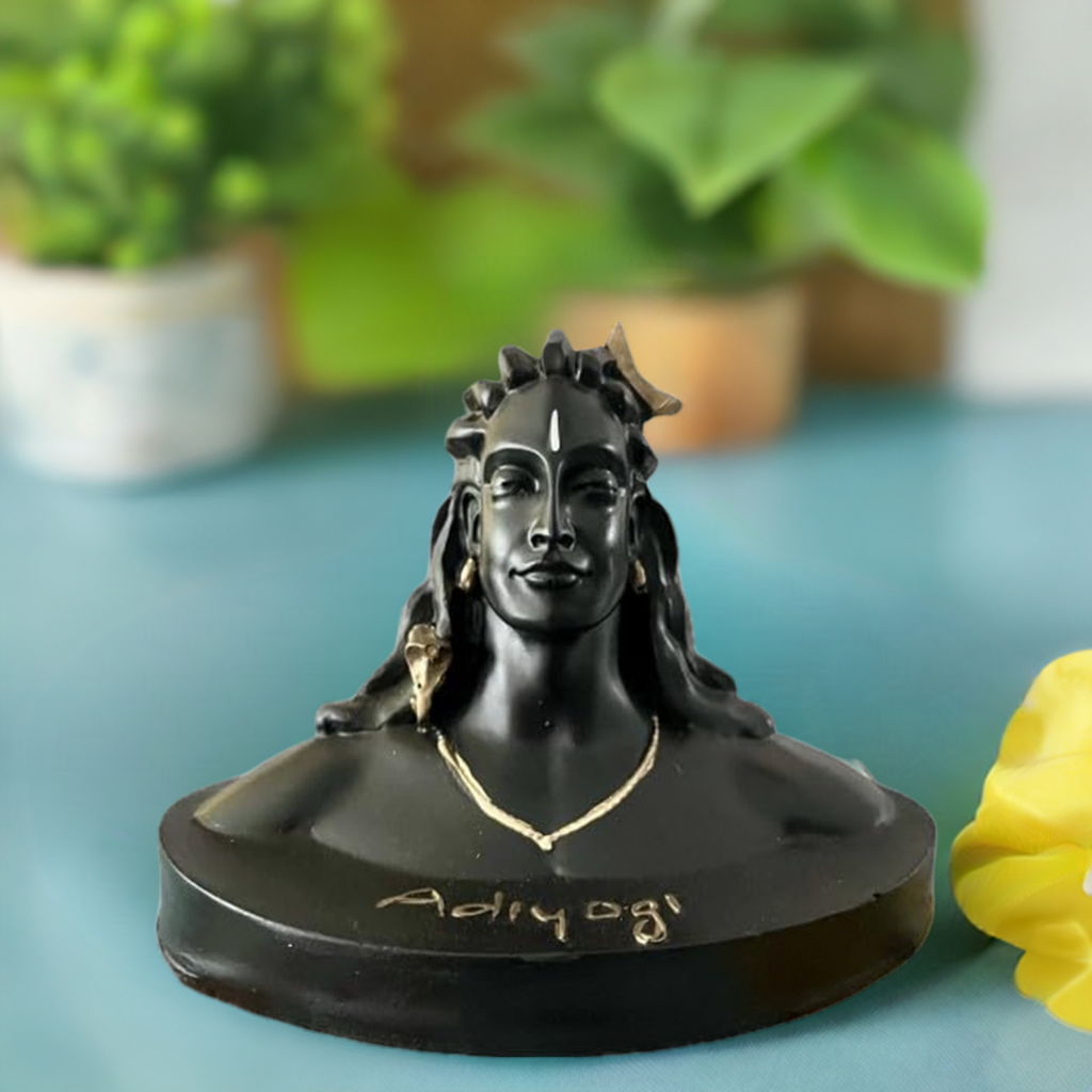 Black bust statue of Adiyogi with detailed facial features, hair styled with a crown, and 'Adiyogi' inscribed at the base, set against a blurred indoor background with plants.