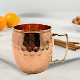 Hammered copper Moscow Mule mug with a brass-colored angular handle on a white surface, with blurred oranges and cinnamon sticks in the background.