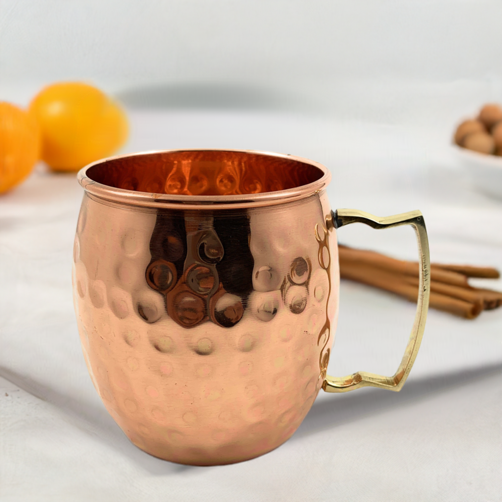 Hammered copper Moscow Mule mug with a brass-colored angular handle on a white surface, with blurred oranges and cinnamon sticks in the background.