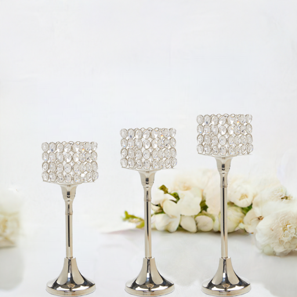 Set of three silver-toned candle holders with crystal-studded square tops in varying heights, placed on a white surface with soft white flowers in the background.