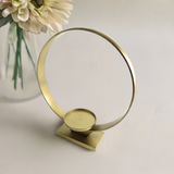 Brushed gold circular candle holder with a flat round base and a square platform, displayed on a white surface with a white flower in a glass vase nearby.